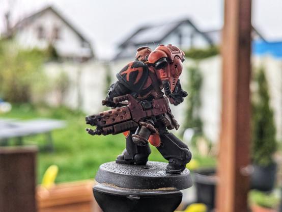 Figurka Space Marine w pomarańczowo-czarnych barwach, widok z tyłu i boku. Wyraźnie widoczne są detale pancerza, plecak oraz uzbrojenie – miotacz płomieni. Na naramiennikach widoczne są pomarańczowe oznaczenia. Tło stanowi rozmyty ogród z domami.