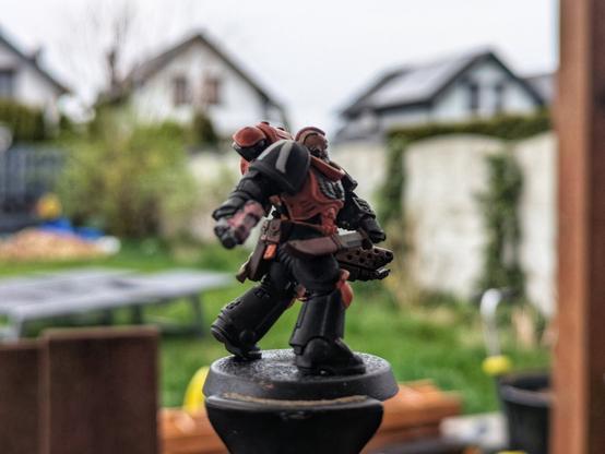 Pomalowana figurka Space Marine w barwach pomarańczowo-czarnych, widok z boku. Dobrze widoczny jest naramiennik z pomarańczowym symbolem oraz piła miecz z licznymi detalami. Figurka stoi na okrągłej podstawce, sfotografowana na tle ogrodu i domów.