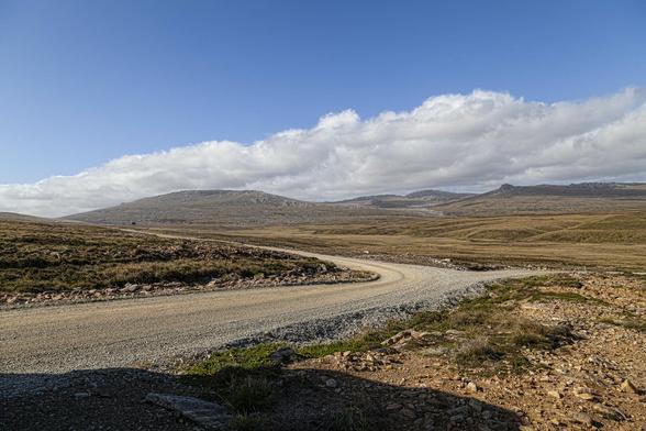 We're off to one side of a rough, narrow road that winds from our left ahead of us to the right then curves back on itself and up over a rise on the left side where it disappears. The landscape here is one of low, gentle hills and it feels very dry. The road is a coarse route of crushed stone and gravel. The land is a mix of sandy-coloured soil and stone with tufts of dry grass, mostly in yellows and browns but with some greenery near the road verges. Overhead the sky is clear blue, feeling cool, but a mass of bubbling white cloud builds over the hilltop line towards the horizon.