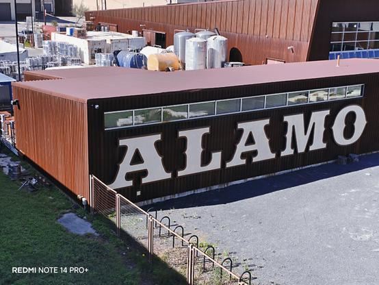 A warehouse with the name Alamo near the Hays Bridge. There is a chain linked fence surrounding the parking lot. There is a bike rack within the parking lot. There are pallets ready to be shipped.