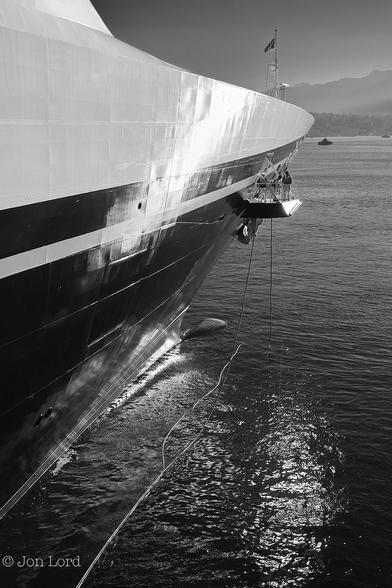 A Black And White Abstract Seascape Photo In Portrait Format. Stretching From The Left Margin Towards The Upper Right Corner Is The Forward Starboard Part Of The Hull Of A Large Ship That Fills Most Of The Frame. The Lower Two-Thirds Of The Hull Is A Glossy Black With The Horizontal Welded Seams Clearly Visible. Above Is A Brilliant Glossy White. Both The Black And White Catching The Still Low Early Morning Sun. On The Bow Is A Small Mast Flying A Flag. Aft Of The Bow On The Upper Black Part Of The Hull Is A Large Hatch That Has Been Hinged Downwards Forming A Platform With Safety Rails Surrounding It. Standing On It Are Three Crew Handling The Lowering Of A Number Of Mooring Lines That Are Visible Hanging Down From The Platform Into The Water And Then Stretching Aft Parallel To The Hulls Waterline. The Water Below Is Jet Black With Some Reflected White Sparkles From The Sun. Beyond A Few km Away Is A Ferry, A Little Larger Than A Speck. In The Background Is A Sloping Mountainous Ridge With A Clear And Cloudless Sky Above. 
The Location Is Vancouver, BC. The Vessel Is The 'Disney Wonder' Cruise Ship (2017).