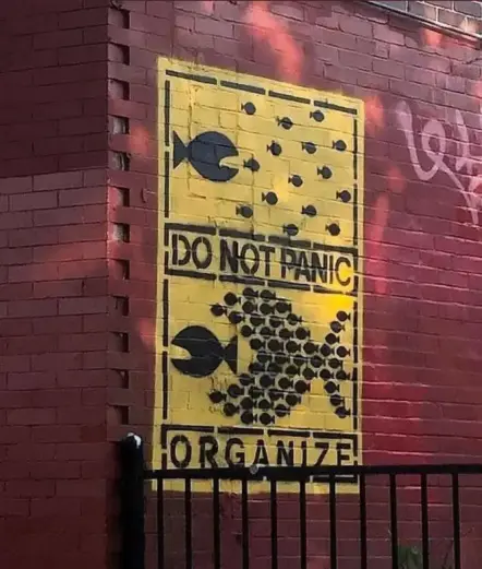 Yellow wall mural shows small black fish uniting to form a larger fish facing off against a predator. Text reads: “DO NOT PANIC / ORGANIZE.”