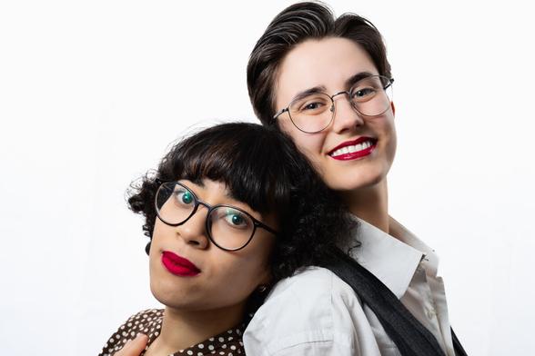 Two individuals stand against a white background. Both wear round glasses and red lipstick. The person on the left has short, dark, curly hair with bangs and tilts their head slightly toward the right. The person on the right has short, neatly styled hair and leans back against the other person’s shoulder, smiling. Both appear relaxed and content.
