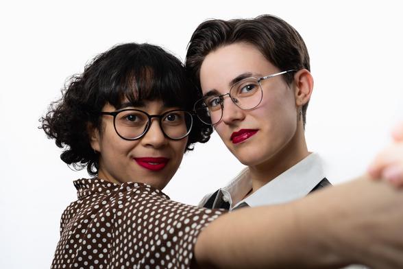 Two people pose close together against a white background. Both wear round glasses and bright red lipstick, looking directly at the camera with relaxed smiles. The person on the left has short, curly black hair with bangs and wears a brown polka-dot blouse. The person on the right has short, neatly combed hair and wears a collared shirt with a dark vest. In a friendly, selfie-like shot, they appear to be extending an arm toward the camera.
