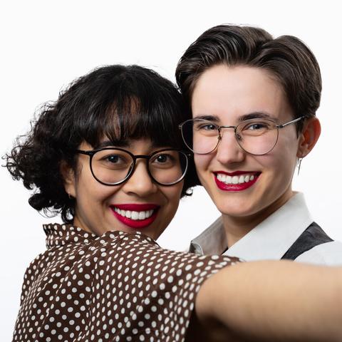 Two people smile at the camera against a white background. Both wear round glasses and bright red lipstick. The person on the left has short, curly hair with bangs and wears a brown, white-dotted shirt. The person on the right has short, neatly combed hair, a white collared shirt, and a dark vest. They appear to hold the camera at arm’s length, leaning close together.