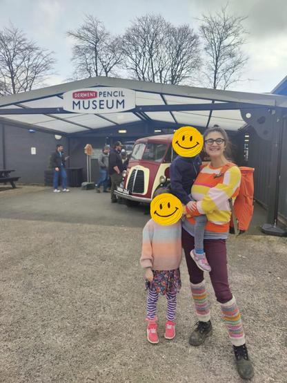 A photo of a mixed heritage woman with dark hair, glasses, an orange backpack, burgundy leggings, grey shoes, striped legwarmers, and a striped jumper in yellow, white and orange. On her hip is a child (dressed in muted colours) and by her feet a slightly older child (dressed in pastels). In the background is a low building and a vintage vehicle and a big sign reading "Derwent Pencil Museum"