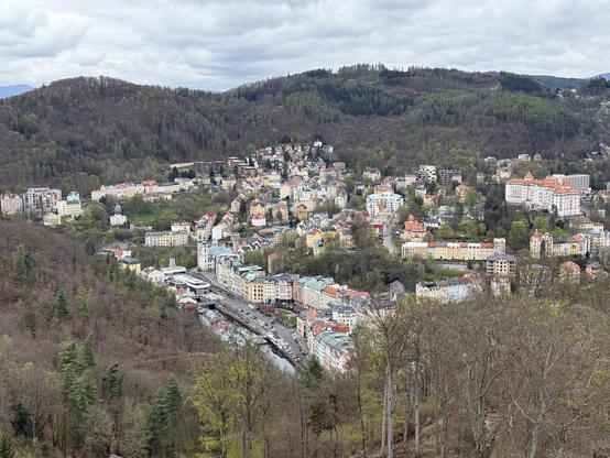 Ein Panoramablick auf eine malerische Stadt, die in einem Tal liegt, umgeben von grünen Hügeln. Die Stadt weist eine Mischung aus farbenfrohen Gebäuden und Strukturen auf, wobei einige historische Bauwerke sichtbar sind. Die Landschaft zeigt eine Mischung aus städtischen und natürlichen Elementen, darunter Bäume und ein Fluss