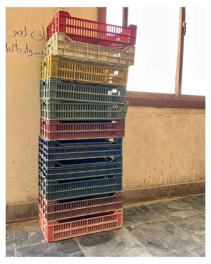 The image shows a stack of colorful plastic crates placed near a window in a room with tiled flooring. The crates are stacked in a vertical alignment, and there are several colors visible, including red, yellow, green, blue, and dark blue. On the wall, above and to the left of the crates, there is Arabic script.