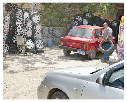 This image shows a man holding a tire next to a red car at a car service or repair place. The background is filled with a large collection of car tires and various styles of car wheel rims. There are some parked cars and an old chair in the scene, suggesting an informal or small-scale automotive service area. The outdoor setting and the items around indicate that this might be a place where tires and rims are sold or replaced.