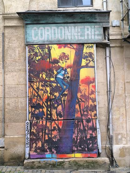 Das Bild zeigt eine Wandmalerei an einer Fassade eines Gebäudes, die eine Szene mit einer Figur darstellt, die auf einem Baum sitzt. Der Hintergrund ist in warmen Farben wie Orange, Rot und Gelb gehalten. Der Baum ist detailliert dargestellt, mit dunklen Silhouetten, die sich gegen den leuchtenden Hintergrund abheben. Die Figur könnte eine Katze sein.