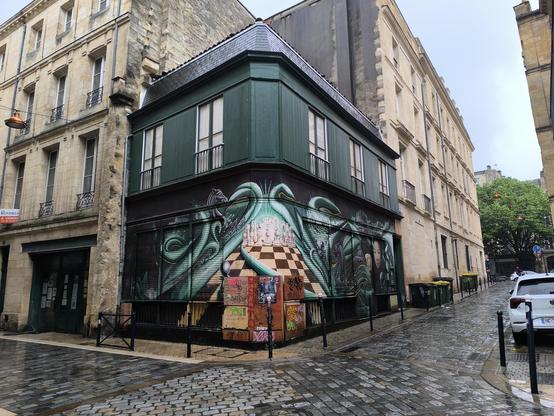 Das Bild zeigt ein Gebäude bzw. Die Ecke davon. Das Gebäude hat eine dunkelgrüne Fassade und befindet sich an einer Ecke, umgeben von weiteren Gebäuden mit Steinmauern und Fenstern. Die Wandmalerei zeigt eine fantastische Szene mit grünen Tentakeln, die über ein Schachbrett mit Figuren streichen. Jeweils rechts und links am Ende des Bildes ist ein menschlicher Kopf gezeichnet.