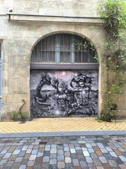 Das Bild zeigt eine beeindruckende Wandkunst auf einer Tür, die in einem Gebäude mit einer steinernen Fassade und einem Bogenfenster mit Gittern integriert ist. Die Wandkunst ist in Graustufen gehalten und zeigt eine surreale Szene mit verschiedenen fantastischen Figuren und Motiven, darunter eine Katze mit großen Augen und Gesichter in den Wolken. Die Szene ist detailliert und kreativ, mit einer Mischung aus organischen und abstrakten Formen.
