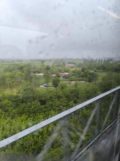 Blick aus dem Zugfenster: wir fahren ziemlich hoch, in der Ferne sieht man viel Grün und ein paar Häuschen. An der Scheibe sind unscharf Regentropfen zu erkennen. Der Himmel ist grau.