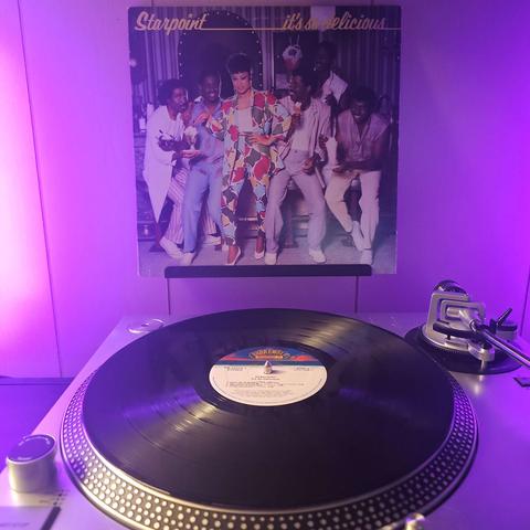 A vinyl record sits on a turntable. Behind the turntable, a vinyl album outer sleeve is displayed. The front cover shows the members of Starpoint in an ice cream shop. The 5 male members holding desserts, while the female member is pointing at one of them to her left.