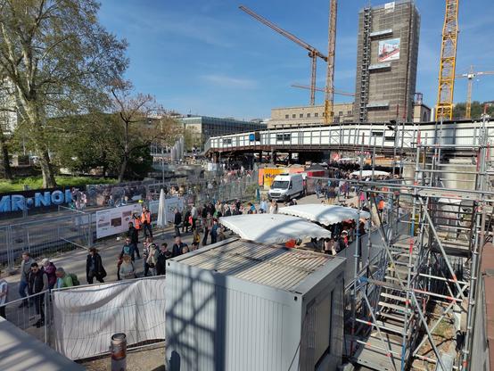 Blick aus erhöhter Position.
Im Vordergrund sind Menschen zwischen Bauzäunen.
Im Hintergrund ist der östliche Fernwanderweg und Turm des Bonatzbau zu sehen.