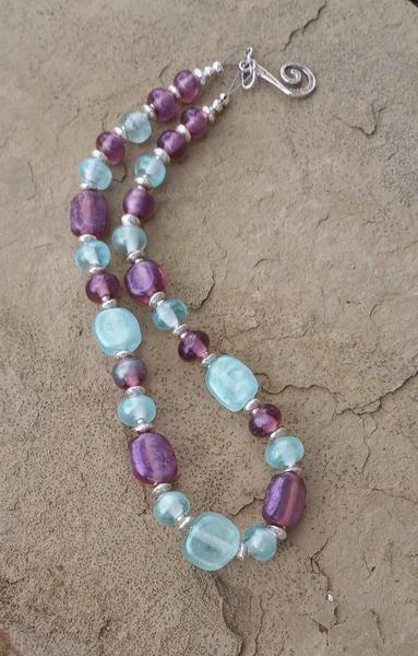 Image of a handmade glass bead necklace in pale blue and amethyst colors set against a beige stone.