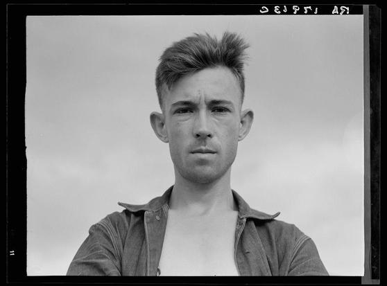 This black and white photograph depicts a young man with disheveled hair standing against an overcast sky. He appears to be wearing a sleeveless undershirt underneath a loosely fastened denim shirt, which is unbuttoned at the collar. His expression is serious as he looks directly into the camera lens.
The image has some text on it: "DEJPSI" in reverse written across the top and bottom edges of the photograph, suggesting that this may be a photo from an archive or collection with reversed orientation. In addition to the main subject, there are no other significant background elements visible; however, faint outlines indicate what could possibly resemble distant trees or structures against the sky.
This image is associated with Dorothea Lange's work on sharecroppers in Macon County, Georgia during a time when individuals received $5 per month from landowners. The specific photo number provided as context for this particular photograph (6495b785ba25398d90c4ea6e.jpg) could be used to find more information about the original source or background story of this image in a database like Sharecropper, which likely contains historical records on sharecroppers and their living conditions.