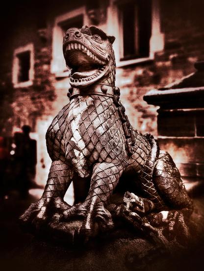 A sepia-toned photograph shows a weathered stone statue of a mythical creature, possibly a dragon or griffin, in a slightly low-angle shot. The creature is depicted with a reptilian head featuring sharp teeth and a long snout pointing upwards and slightly to the right. Its body is covered in scale-like patterns, and it has strong, clawed paws resting on a rocky base. A thick, studded collar encircles its neck, and chains appear to drape over its body. The statue is positioned outdoors, with blurred architectural elements of a building with windows visible in the background. To the left, a dark, indistinct figure of a person can be seen. The overall atmosphere is somewhat gothic and aged, emphasised by the monochromatic tones and the worn appearance of the stone.