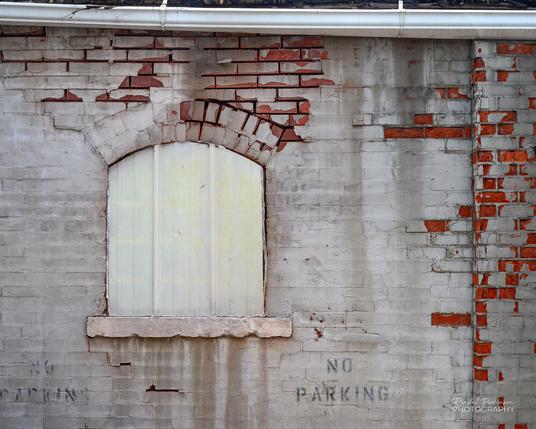 A brick wall with peeling white paint has a window closed off by metal siding.  "NO PARKING" is painted in two locations on the wall.