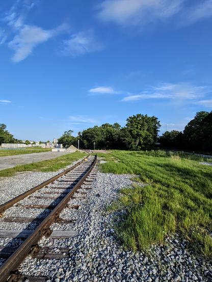An old set of rusty train tracks with weathered wooden ties, on a bed of grey gravel lead through a green grassy field to an old industrial plant set far back from the road, where a variety of buildings and silos appear in the distance beneath  big blue Florida skies.