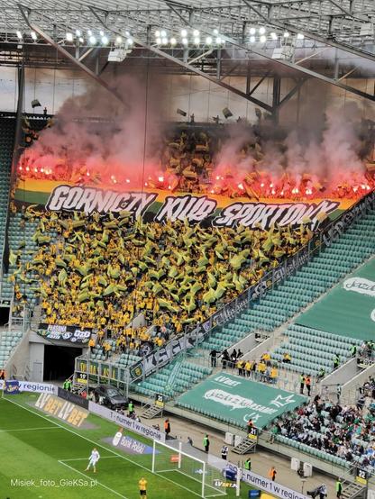 Zdjęcie przedstawia trybunę stadionu piłkarskiego wypełnioną kibicami GKS Katowice. Fani ubrani są głównie w żółte koszulki, trzymają flagi w barwach klubu (żółto-zielono-czarne), a nad ich głowami rozciągnięty jest transparent z napisem „Górniczy Klub Sportowy”. W tle unoszą się kłęby czerwonego dymu z rac odpalonych przez kibiców. Trybuna jest niemal w całości zapełniona, a atmosfera na zdjęciu jest bardzo intensywna i pełna emocji. W dolnej części kadru widoczny fragment boiska i bramkarz.