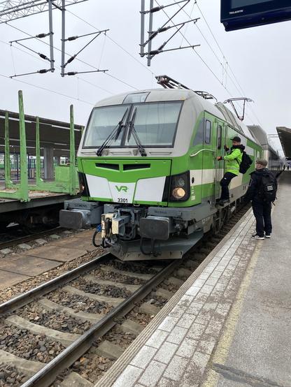 Kuljettaja kiipeää asemalaiturilta sähköveturin ovelle. Toinen mies seuraa vierestä. Veturin perässä on kaksikerrosmatkustajavaunuja, viereisellä raiteella tyhjiä puutavaravaunuja.