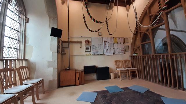 Cushioned chairs on the left against a large glazed perpendicular window with clear glass. Bell ropes with sallies suspended from the ceiling. Notice boards on the far wall. Glazed oak screen on the right overlooking the church.