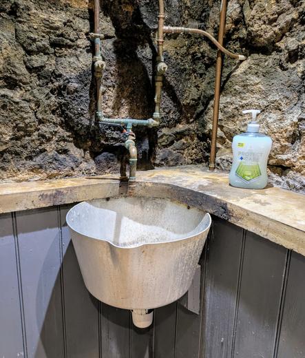 A corner view of a room. The top half of the walls are rough stone, with painted wood panelling underneath and a wooden ledge dividing the two. There are bare copper pipes against the walls and a white sink that looks like the top half of a bucket stuck in the corner under the wooden ledge.