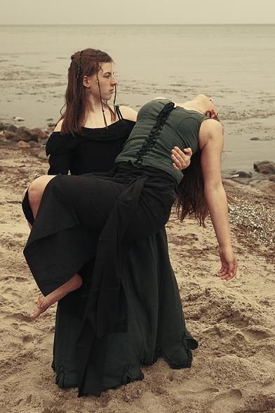 Ein Mädchen trägt am Strand ein anderes bewusstloses Mädchen. Bei dem Mädchen, das getragen wird, ist an der Seite des Kopfes Blut zu erkennen. Beide tragen eher altertümliche Kleidung.