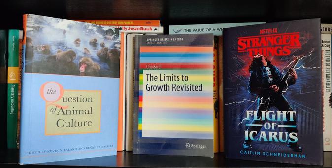 A photo of three books standing on a black shelf against a background of other books. On the left, The Question of Animal Culture, showing Japanese macaques bathing in a hot spring. In the middle, The Limits to Growth Revisited, showing the title on a background of coloured horizontal bars. On the right, Stranger Things: Flight of Icarus, showing a drawing of a young man, Eddie Munson, rocking out on a black Flying V electric guitar with lightning bolts shooting off the guitar at both ends.