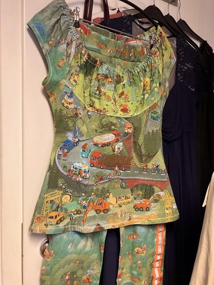 An einem Kleiderbügel an einer Wand hängt eine bunte Kombination aus kurzärmeligem Oberteil mit großem, gerafften Ausschnitt. Die Hose ist geschnitten wie eine Leggins und hat einen orange-reflektierenden Streifen an den Seiten. Der Stoff ist bedruckt mit Motiven wie aus einem Wimmelbildbuch von Ali Mitgutsch. 
Es ist eine kurvige Straße in einer bergigen Landschaft zu sehen, Autos, Menschen, Kühe, ein Eiswagen, Baufahrzeuge, Rohre, Rennradfahrer, ein Holzlaster… Auf der Hose ist eine Strandszene, mit badenden Kindern, Schlauchbooten, einem Wasserball.