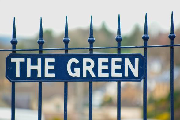 A photo in landscape format, of a set of railings with a street name sign on it; the background is out-of-focus. The street is called 'The Green'; the railings and sign background are painted blue, and the lettering is white.