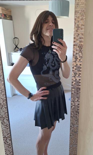 Non binary person in a sheer top with a dragon motif and black leather style pleated skirt