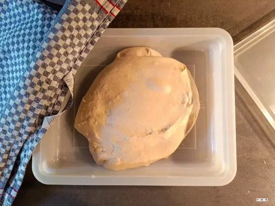 In einer geölten Wanne ruht ein Brötchenteig. Am linken Rand der Wanne liegt ein kariertes Grubentuch, am rechten der Deckel.