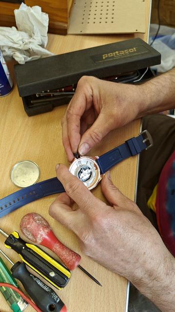 A fixer holds watch and tries to re-attach the crown that has fallen out. The back of the watch has been removed and you can see the internals of the watch.