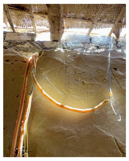 The image shows a setting that appears to be inside a rustic building or a structure, possibly a hut or a traditional dwelling. Key features of the image include:

1. **Structure of the Building**: The roof is made of thatch, indicating a rustic or traditional construction style. Thatch roofing is often used in rural or traditional architectures and offers an ecological and insulating roof solution. The poles supporting the roof and the coarse masonry walls also hint at a simple construction method.

2. **Electrical Installation**: The image prominently features an arrangement of electrical conduits and wires within a transparent flexible tubing, which is zigzagging from the roof area and extends towards the ground. This setup suggests a provisional or makeshift electrical installation, possibly for temporary lighting or power supply.

3. **Textural Details**: The walls and the ground are textured with what appears to be a dark sand or fine dirt, a characteristic that might indicate the building is located in a coastal or desert region where such building materials are prevalent.

This location might be used for various purposes—residential, community, or possibly even for some small-scale industrial activities considering the apparent electricity rig setup.