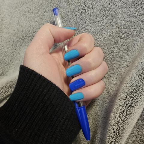 Photo de ma main qui tient un bic bleu. Tous les ongles bleu lagon sauf l'annulaire bleu roi comme le capuchon du bic. L'ensemble en fini glossy