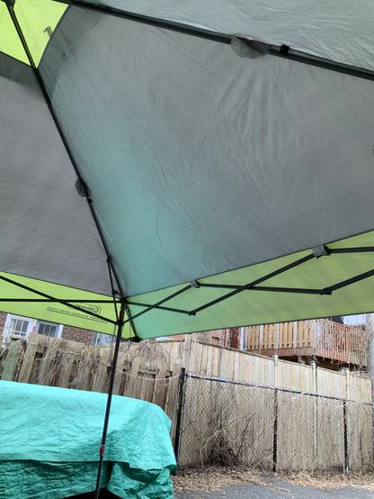 View of the inside roof of a green and green instant shelter set up in a back yard with a metal and wooden fence and balconies in background, as well as a green tarp covered stuff!