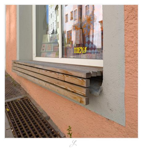 The picture shows a windowsill of a store that is completely covered by a simple wooden bench. It was attached directly to the windowsill. The bench consists of nine or ten rectangular wooden bars, which are mounted in such a way that they form a right angle on which you can sit. There are paper cards in the window, and the word “new” is written in yellow curved letters on the window. The window reflects the buildings on the opposite side of the street and a thin vertical line of blue sky.

AI disclaimer: Using my work, its meta data, written or derived description to create media with or train AI based systems is prohibited.