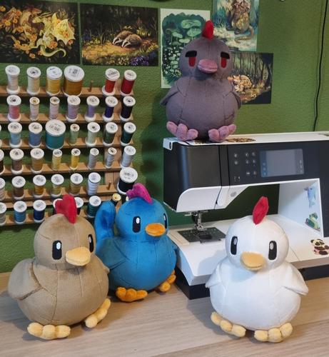 Quatre peluches de poules inspirées de celles du jeu Stardew Valley, de gauche à droite la brune, la bleue, la grise et la blanche. Elles sont posées sur un bureau et la grise est posée sur une machine à coudre.