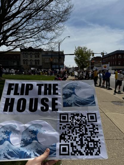 A hand holds a sign that reads "FLIP THE HOUSE," accompanied by images of ocean waves and a QR code. In the background, a group of people is gathered, likely participating in a protest or rally. The setting appears to be an outdoor