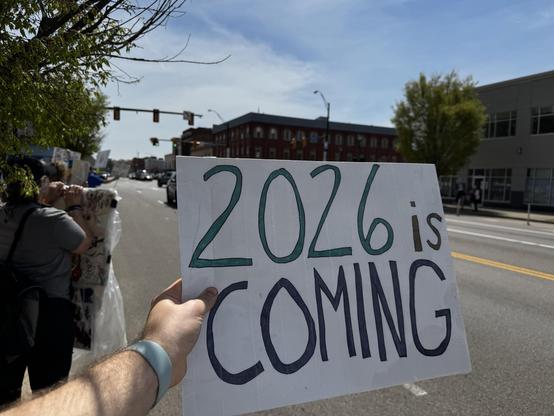 A person holds a sign that reads "2026 is COMING" in large letters. The background shows a street with traffic light and some buildings, while others nearby appear to be participating in a gathering or protest. The scene is bright and captures a