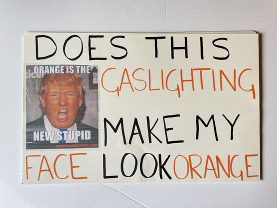 A protest sign has a picture of Trump’s orange face and says, “Does This Gaslighting Make My Face Look Orange”.