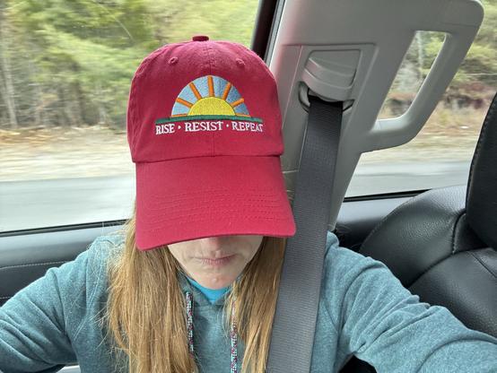A woman takes a selfie in a car to show her hat, a red baseball cap embroidered with a sunrise logo and the slogan, “Rise Resist Repeat,” a call to daily action against the advance of authoritarianism in the US.