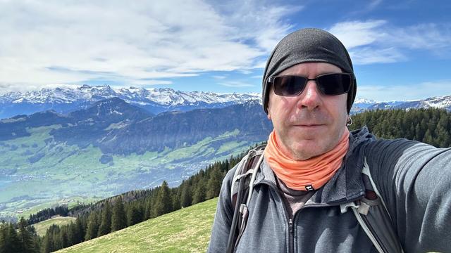 Selfie of me, a middle-aged white guy wearing sun glasses and a gray slouchy beanie with a green valley and snowy Swiss Alps in the background.