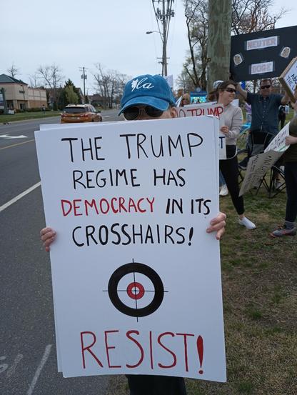 A woman in a blue hat stands by the roadside with a protest sign.  The sign reads:  "The Trump regime has Democracy in its crosshairs!  Resist!"  There is also a drawing of a target with crosshairs.