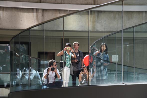 five or six people stand in a group in a walk way with a glass wall. All of them are most of them are holding cameras/smart phones