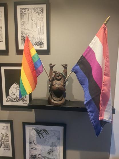 Two flags, a rainbow flag and a gender-fluid flag, are displayed on a light wooden shelf, with a statue of a happy Buddha in the middle holding onto the flags!