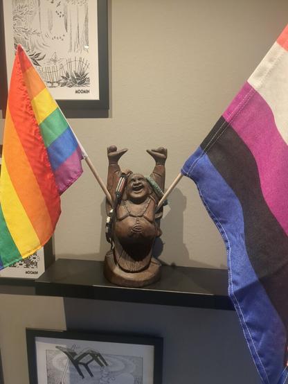 Two flags, a rainbow flag and a gender-fluid flag, are displayed on a light wooden shelf, with a statue of a happy Buddha in the middle holding onto the flags!