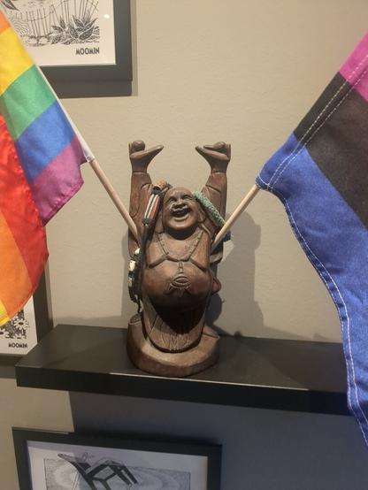 Two flags, a rainbow flag and a gender-fluid flag, are displayed on a light wooden shelf, with a statue of a happy Buddha in the middle holding onto the flags!